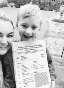 Black and white photo of Chloe and her son smiling at the camera as she holds her dissertation