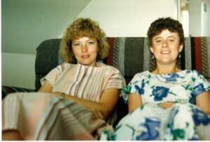 Elaine and a friend sat on the sofa. Elaine is sat on the left and it wearing a beige/brown striped dress. Her friend is sat to the right with short brown hair and a white dress with blue flowers