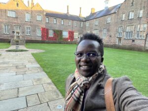 Evan is wearing glasses, a brown coat, beige tartan scarf and brown bag. They are standing in the Quad during the autumn. The leaves in the Quad are an autumnal red