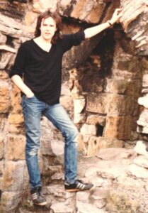 Sean in the late 1980s standing on some rocks. He has long brown hair and is wearing a black long sleeved shirt and blue jeans