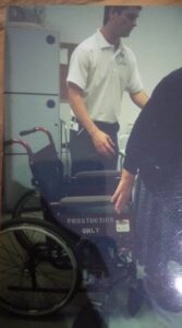 Photograph of Anthony as a student working in practice. Anthony is wearing a white shirt and dark trousers and standing next to a wheelchair.