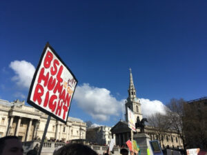 Trafalgar Square, sunny blue sky, on left hand side a protest sign that reads 'BSL is our Human Right'