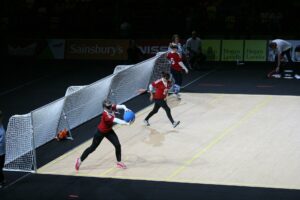 An image of a Goalball game. 3 people dressed in sports wear, each with a blindfold on in front of a large goal. One of the players holds a large blue ball. 