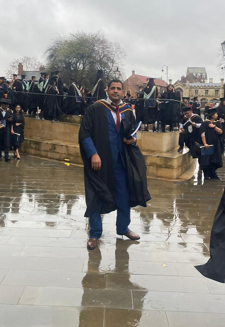 Ahmed is standing outside of York Minster. Ahmed is wearing a black graduation gown and is carrying a black graduation cap and a graduation programme in his hand. Ahmed is wearing a blue suit, white shirt and red tie.