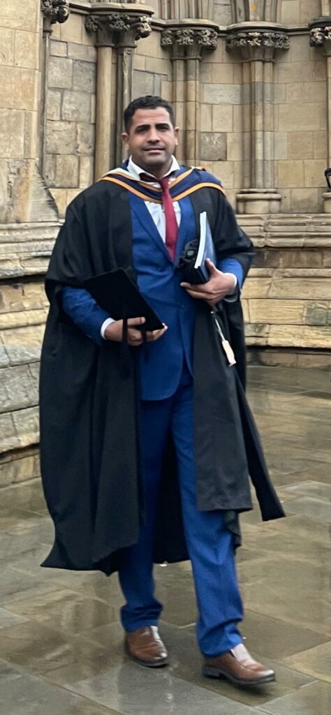 Ahmed standing outside York Minster after his graduation. Ahmed is wearing the York St John chevrons, black graduation gown and is holding his black graduation cap. Ahmed is wearing a blue suit, white shirt and red tie.