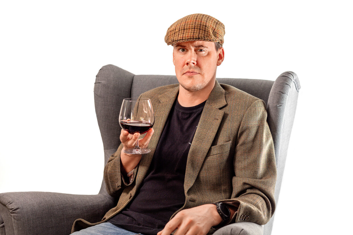 Alex is sitting in an armchair holding a wine glass. He is wearing a flat cap, tweed blazer and black top.