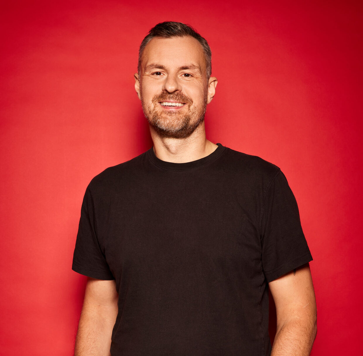Man with short hair and a beard smiling at a camera. The man is wearing a black t-shirt and is standing against a red background