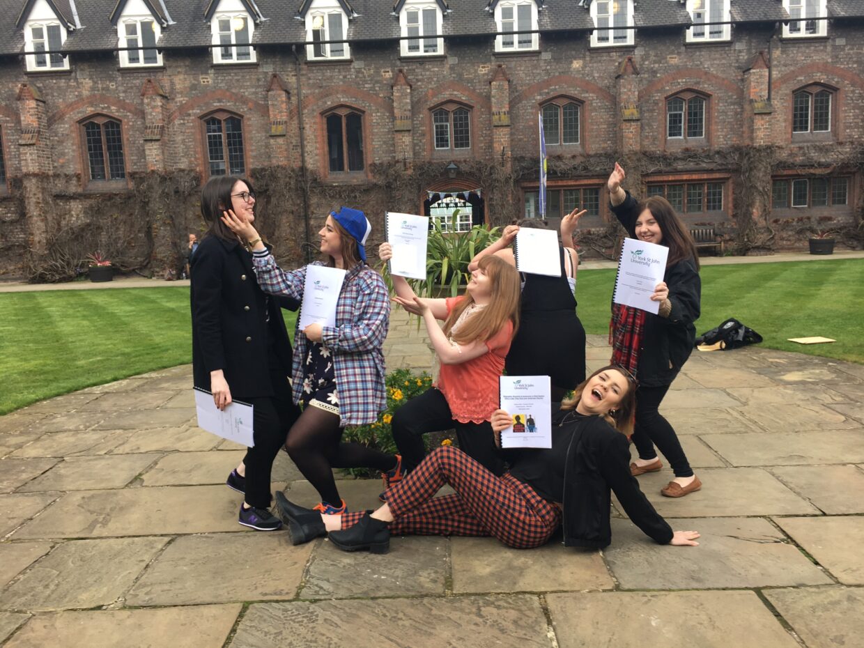A group of students standing in the Quad. 6 people are holding their dissertations and posing in various ways.
