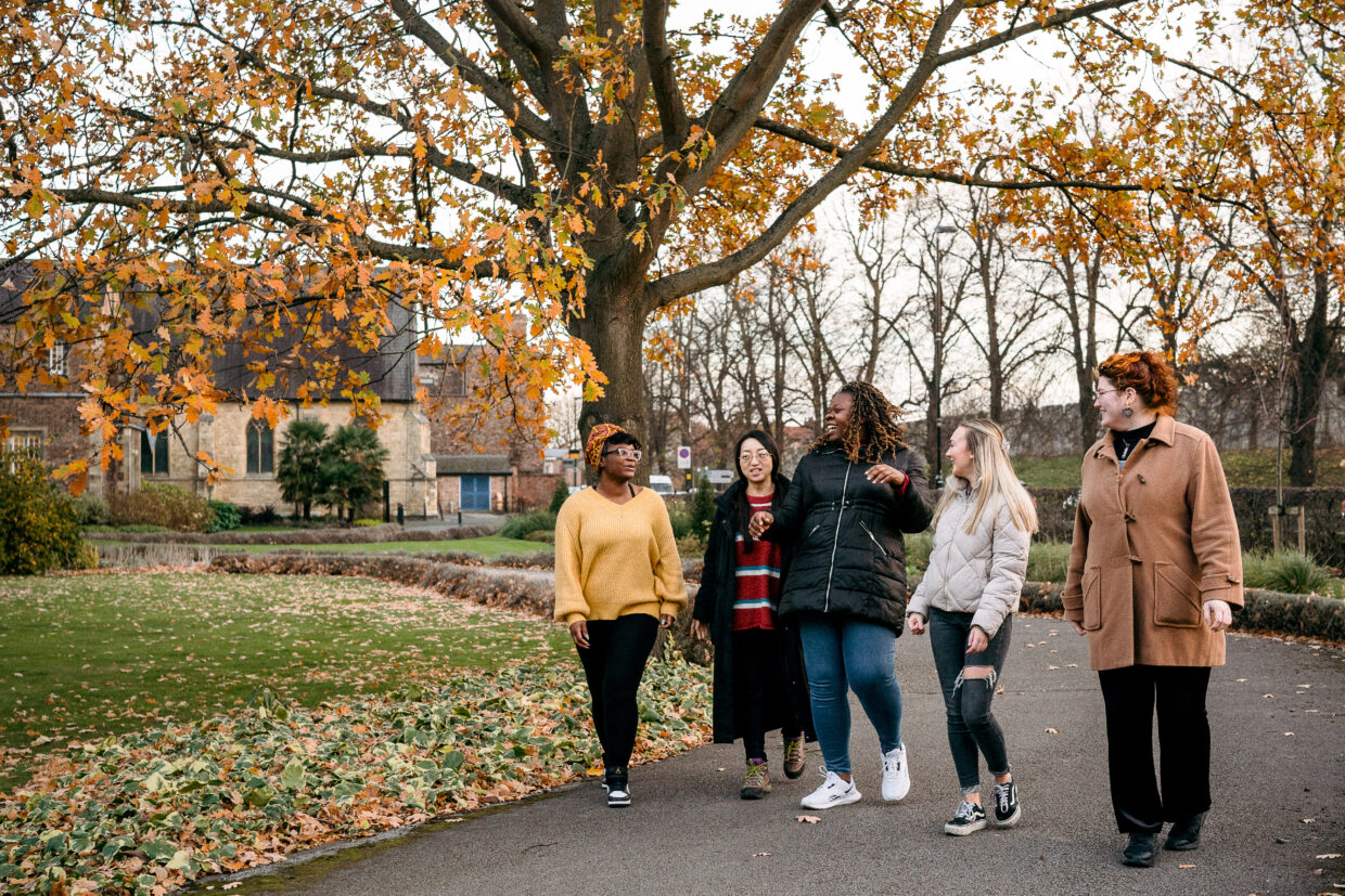 Five university students are walking on the path by the front of Lord Mayors Walk in Autumn. The student on the left is wearing a yellow jumper and black jeans. The student on their right is wearing a black coat over a red jumper with white and blue stripes. The students in the middle is laughing and wearing a black coat and blue jeans. The student on their right is wearing a light grey puffer coat with black ripped jeans on. The final student on their right is wearing a brown coat and black trousers.