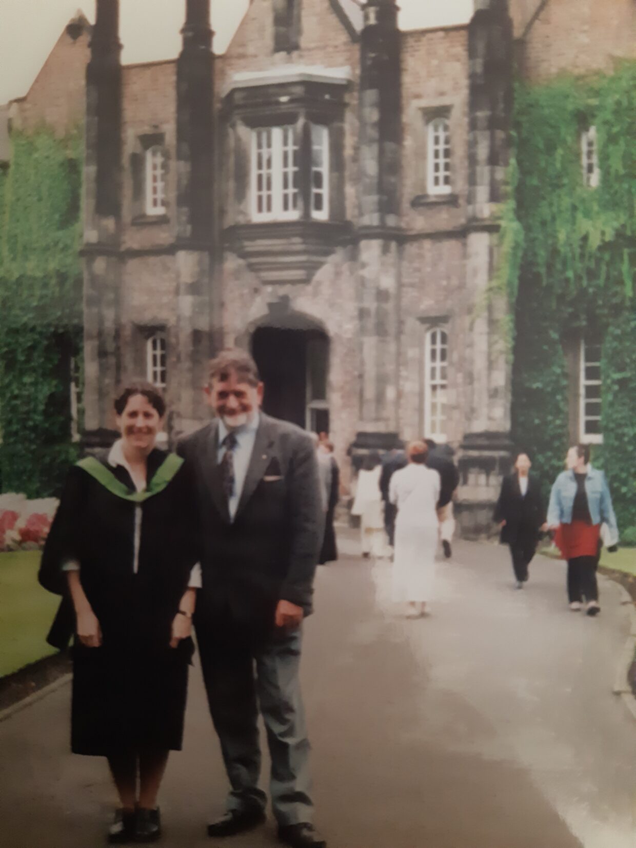 Jennifer is wearing a graduation gown and standing outside of the Lord Mayor's Walk entrance. She is standing with her father who is wearing a suit.