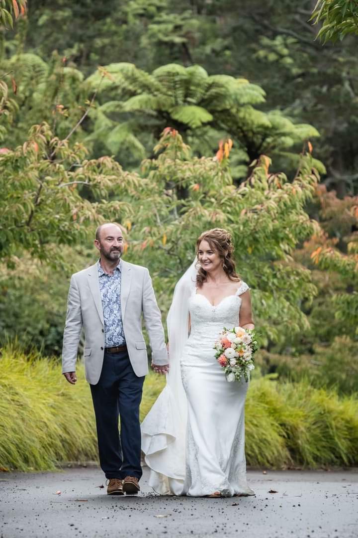 Jeremy is wearing a beige jacket, pale blue shirt and dark grey trousers. He is walking with his wife on their wedding day. His wife is wearing a white wedding dress and veil.