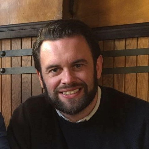 Mark has short brown hair and a beard and is smiling at the camera. He is wearing a black jumper with a white shirt underneath.