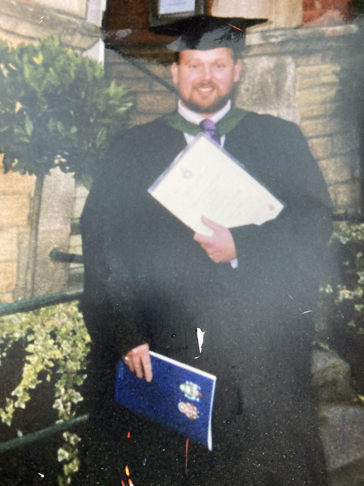Michael is wearing a graduation cap and gown and is holding his degree certificate. He is standing outside in a garden.