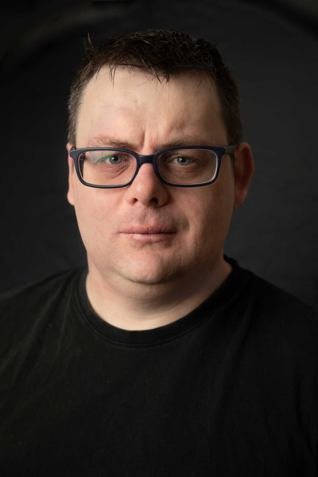 A promotion shot of Pete. Pete is wearing glasses and a black tshirt. He has short black hair and is standing against a black background.