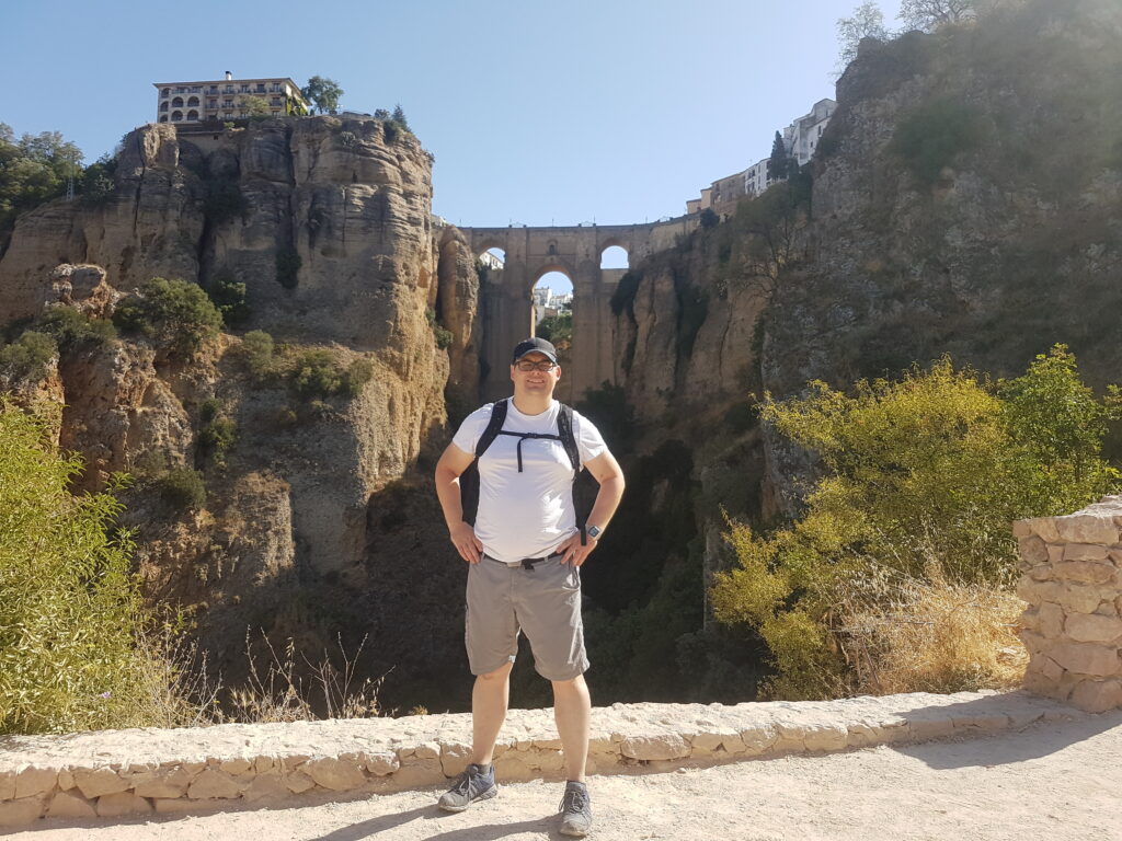 Pete standing in front of the bridge over the gorge at Ronda. Pete is wearing glasses, a cap, white t-shirt and beige shorts.
