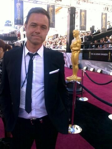 Peter is stood on the Oscar's red carpet and is wearing a black suit, black tie and white shirt. He has short hair and is smiling at the camera.