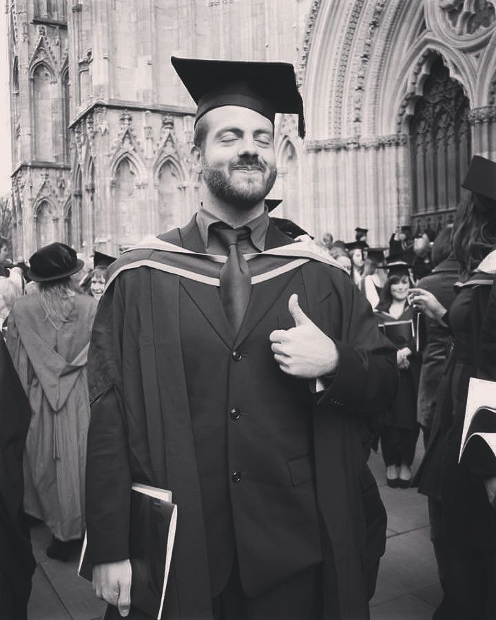 Black and white picture of Riccardo outside of York Minster. Riccardo is wearing a graduation cap and gown, smiling with his eyes closed and with his thumbs up.