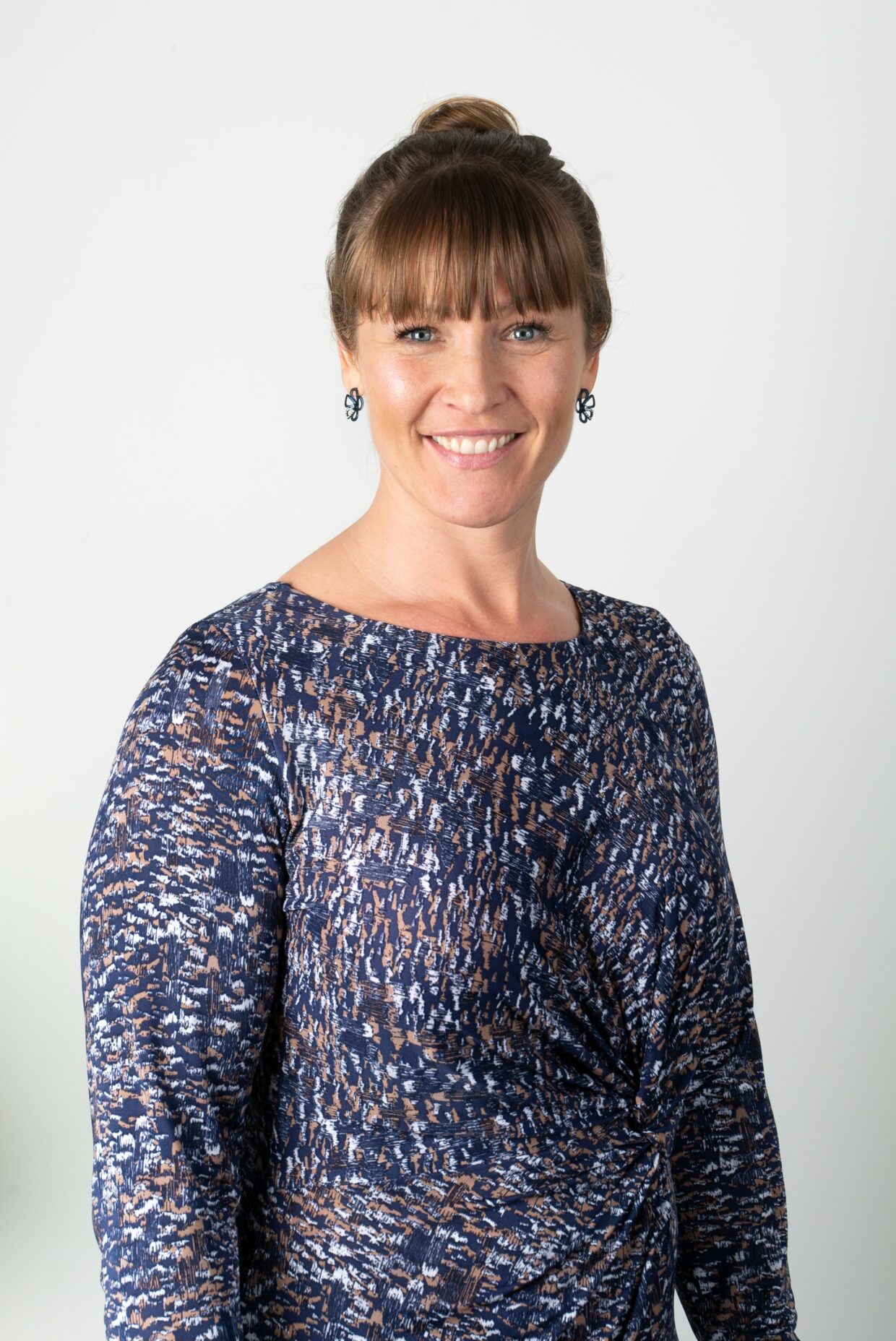 Tracy has brown hair tied in a bun and is smiling at the camera. Tracy is standing against a white background and it wearing a blue top with white and orange dots.