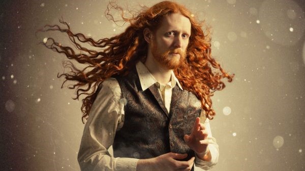 Alasdair has long red hair and a beard. He is wearing a white shirt and brown waistcoat