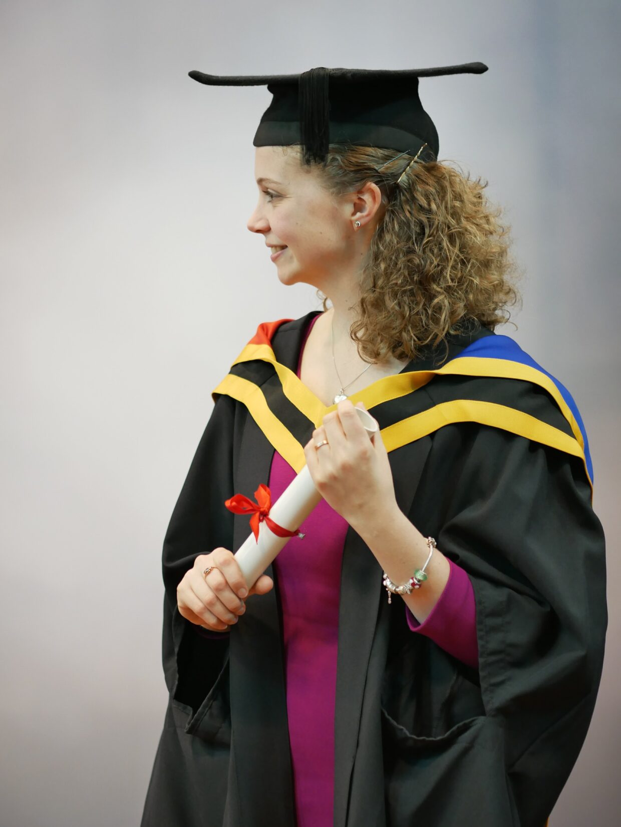 Chloe is stood against a photography background for her Masters graduation photo. Chloe is wearing a graduation cap and gown. Chloe is also wearing a pink dress and holding a scroll.