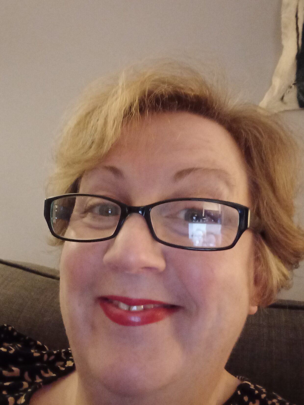Selfie of Elaine smiling at the camera. Elaine has short blonde hair, is wearing square, black-framed glasses and red lipstick.