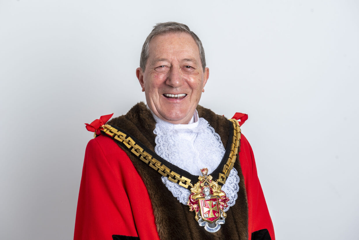Michael has short grey hair and is smiling at the camera. Michael wears his mayoral robes, which are red with black lining and a civic regalia which shows the Wolverhampton coat of arms on a black and gold chevron.