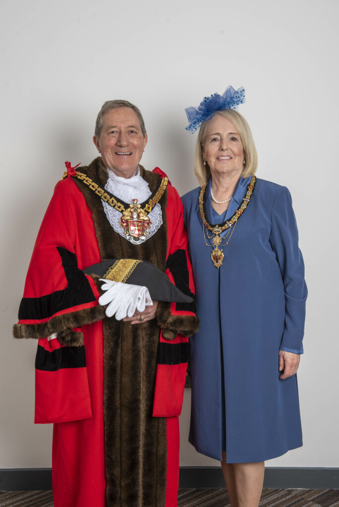 Cllr Dr Michael Hardacre and Mayoress Ms. Lynn Plant. Michael has short grey hair and is smiling at the camera. Michael is wearing red civic robes and a civic regalia showing Wolverhampton's coat of arms. Michael is also holding black and gold civic headwear and white gloves. To Michael's right is Lynn Plant who has short blond hair and is smiling at the camera. Lynn is wearing a blue hat, blue dress, and blue dress coat. Lynn is also wearing a civic regalia around her neck. 