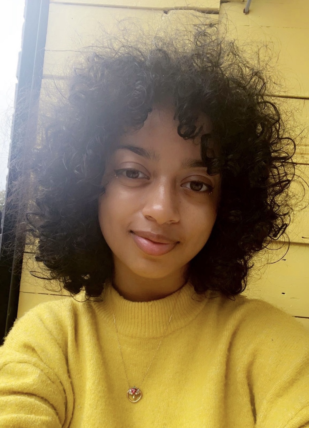 Alisha has brown, shoulder length curly hair and is smiling at the camera. Alisha is wearing a yellow long sleeved turtleneck.