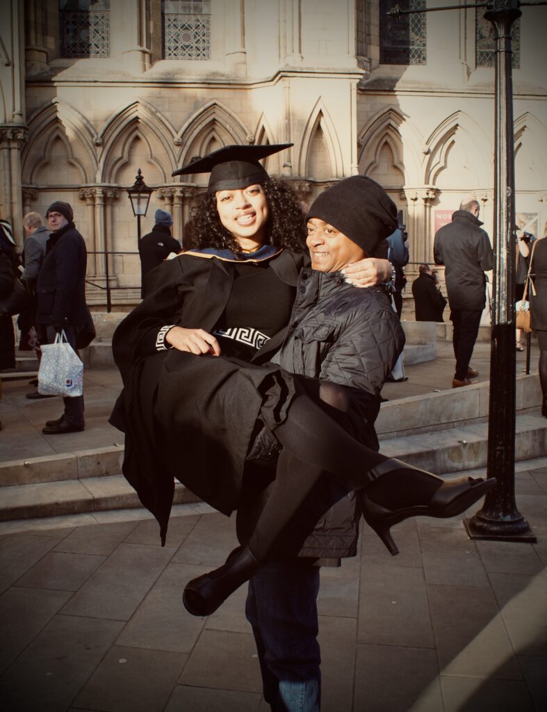 Alisha is in a graduation cap and gown outside of York Minster. Alisha is being picked up by someone in a black coat and hat as they celebrate Alisha's graduation. Both are smiling.