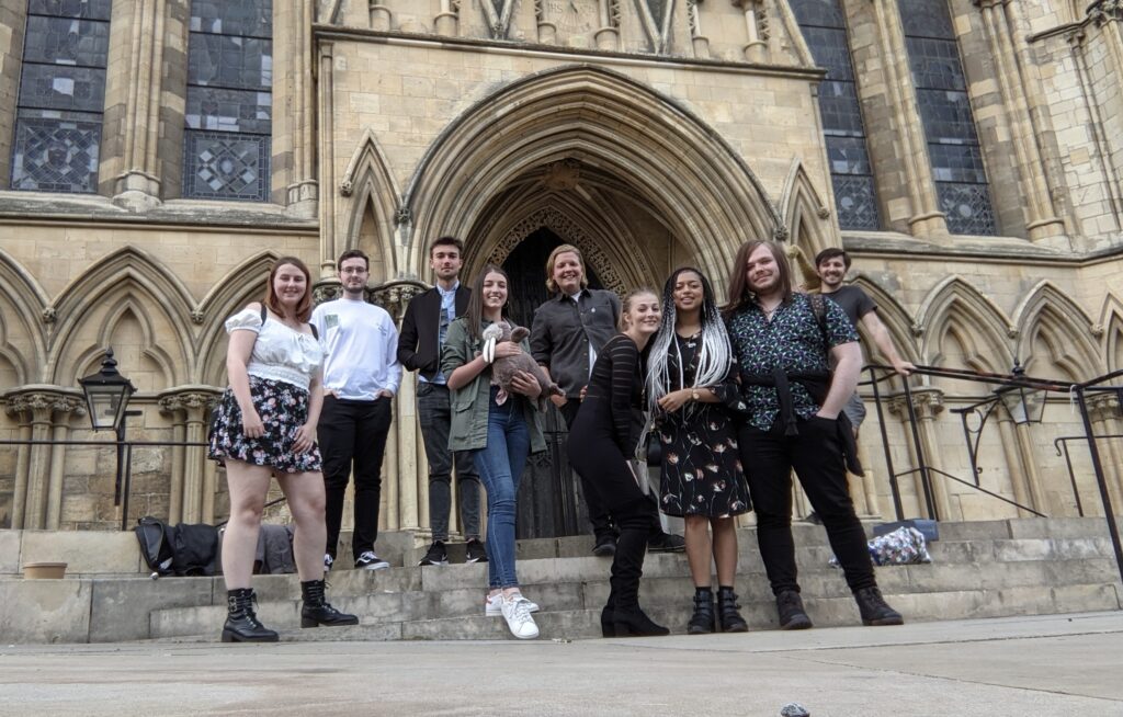 The Animation cohort outside York Minster. 9 students are stood on the steps outside the Minster and smiling at the camera. Alisha is third from the right and is wearing a black patterned dress and black boots.