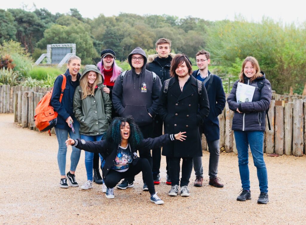 The Animation cohort outside on a trip. It has recently rained as the group are all wearing waterproof coats. Alisha is crouched on the floor with her arms spread widely towards the camera. 