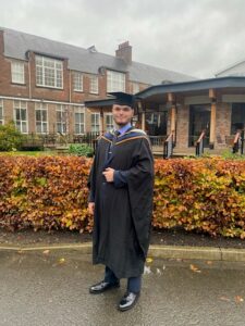 Connor is stood outside the Holgate dining room extension on his graduation day. Connor is wearing a cap and gown and a suit. The weather is overcast and the trees are turning orange for autumn.