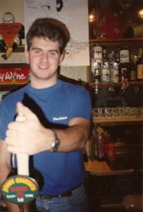 Duncan is behind the bar the the Students' Union pulling a pint of Magners. Duncan has short brown hair and wearing a blue t-shirt and blue jeans