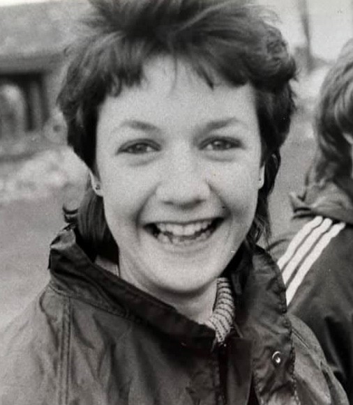 A black and white picture of Heather in her first year of university. Heather has short hair and is smiling widely at the camera. She is wearing a waterproof coat and a light coloured turtleneck underneath.