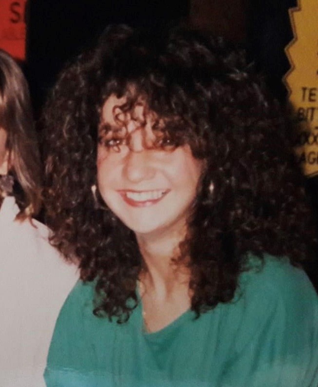 A colour photograph of Heather in her 3rd year of university. Heather has long brown curly hair and is wearing gold hoop earrings and a green t-shirt.