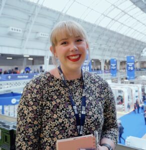 Jenna is at a work event and is smiling at the camera. Jenna has blonde hair tied up in a pony tail. She is wearing a lanyard, navy and cream floral dress and red lipstick. She is holding a pale pink notebook.