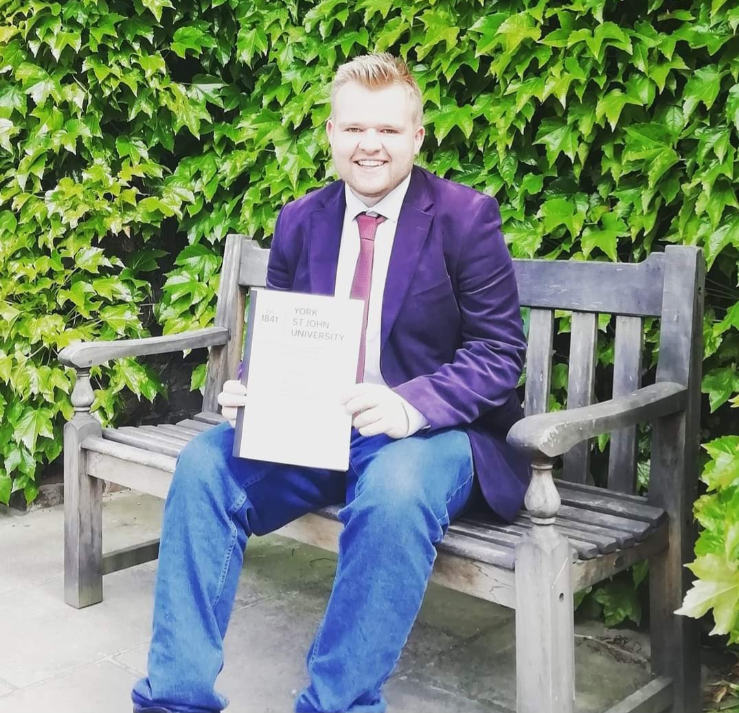 Marc is sat on a bench in the Quad holding their dissertation. Marc is wearing a navy blazer, white shirt, red tie and blue jeans. Marc has short blonde hair and is smiling at the camera.