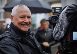 Photo of Martin standing next to a camera in the rain. Martin has short, white hair and is wearing all black. He is smiling at the camera while he prepare to film a segment for BBC News.
