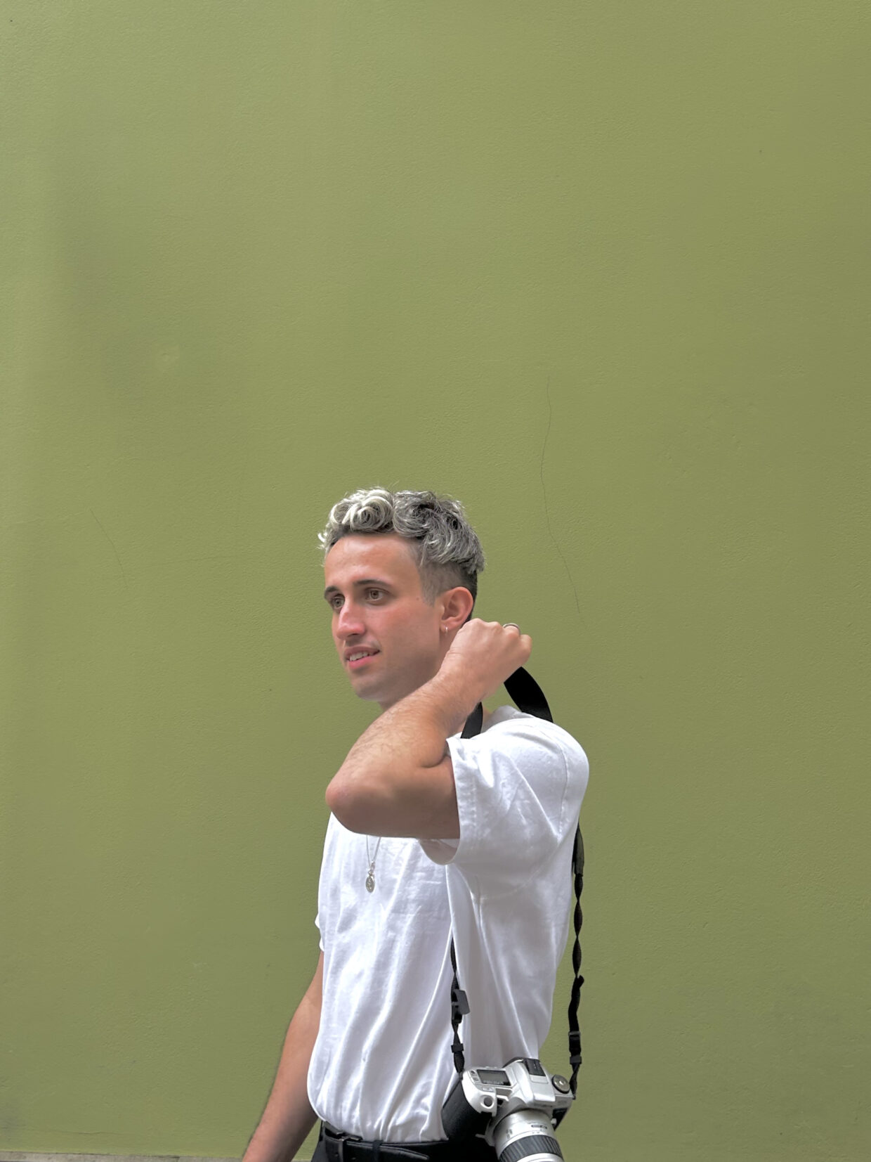 Declan has short silver hair and is wearing a white t-shirt and a camera. Declan is standing against a green background in Poland.