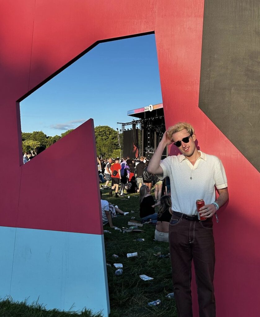 Declan is standing next to the Radio 1 sign at Big Weekend. Declan has short blonde hair, is wearing sunglasses, white short-sleeved shirt and brown trousers.