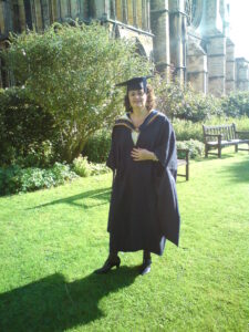 Jennie is wearing graduation robes outside a cathedral after graduating from Huddersfield University. Jennie has short brown curly hair.