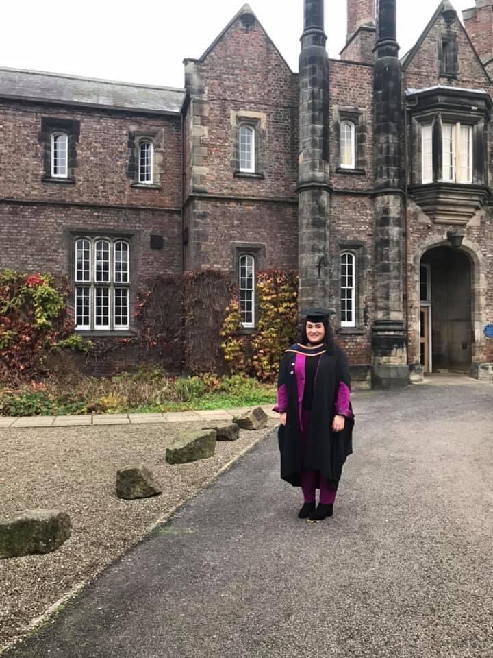 Nicoletta is wearing graduation robes and a purple dress and standing outside the Lord Mayor's Walk building.