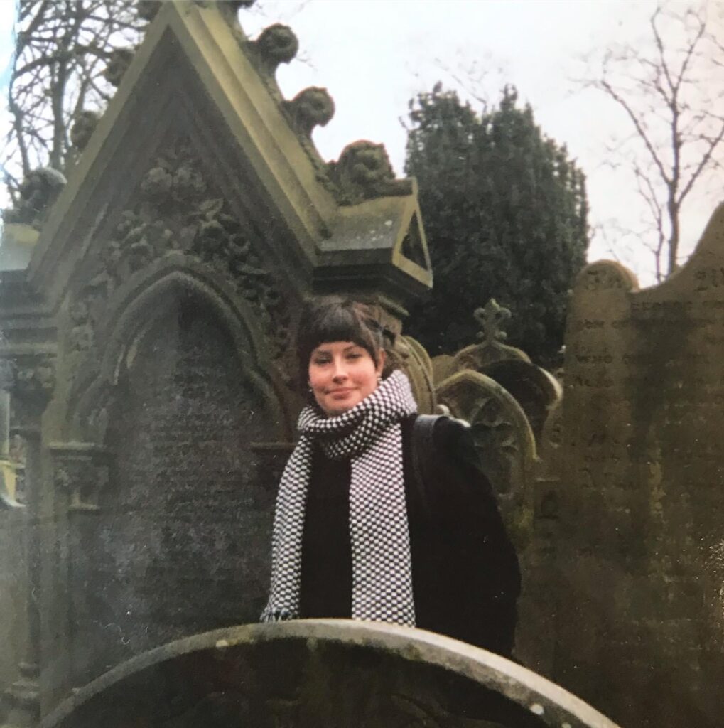 A picture of Tasmin in a cemetery. Tasmin has short brown hair and is wearing a long black and white scarf.