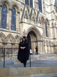 Dana is wearing graduation attire and is stood outside York Minster.