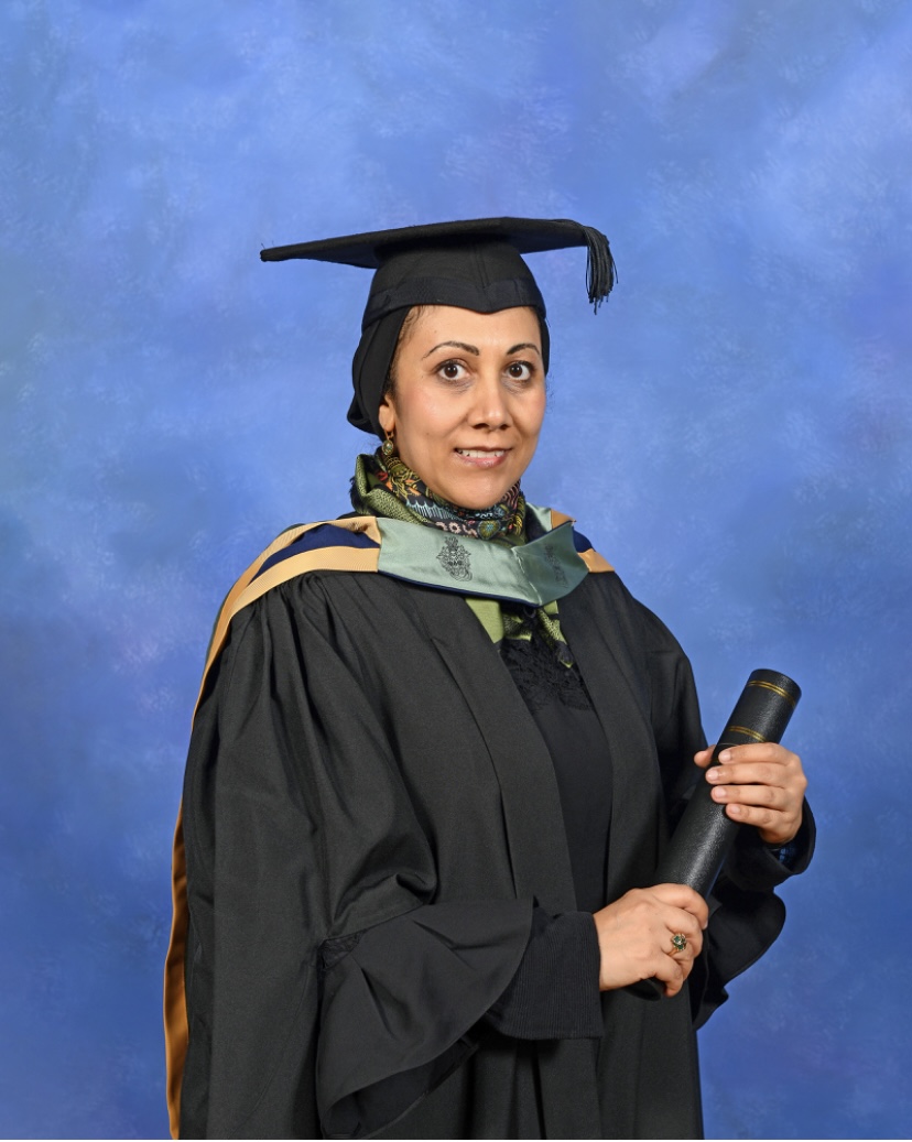 A photo of Mona on graduation day. Mona is standing against a blue background and is wearing a graduation cap and gown.