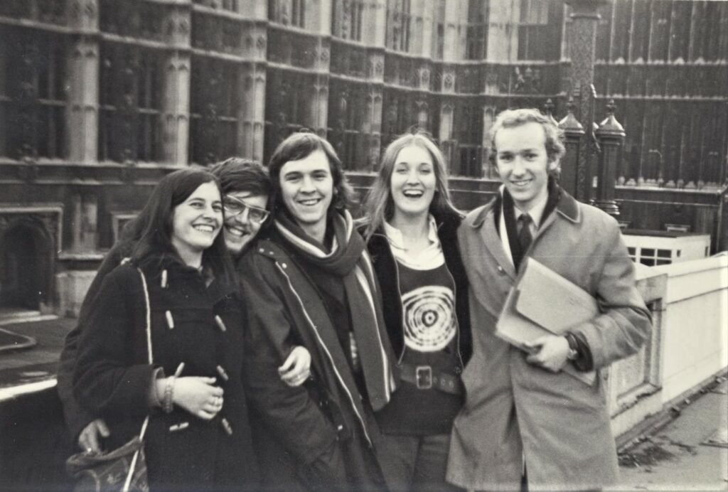 Black and white photograph of five Ripon students in the early 1970s outside of the Houses of Parliament. John is on the far right wearing a light long coat, white shirt and a dark tie.