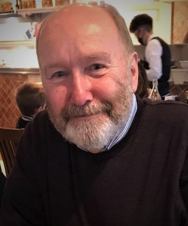 Dr John Ridley has short hair and a short greying beard. John is wearing a dark coloured jumper and and light blue shirt underneath