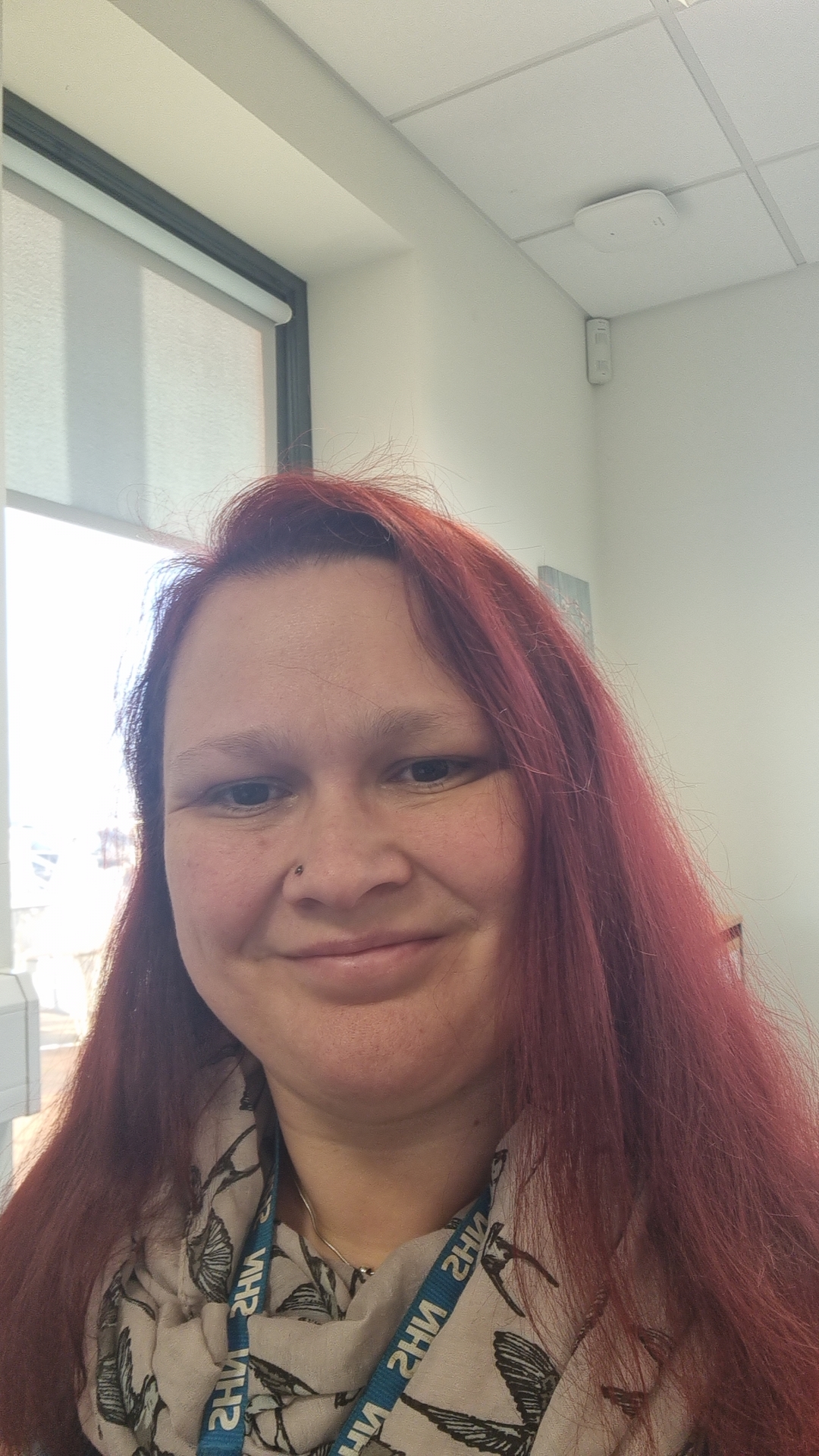 Susan has long red hair and is smiling at the camera. Susan wears a light colour scarf.