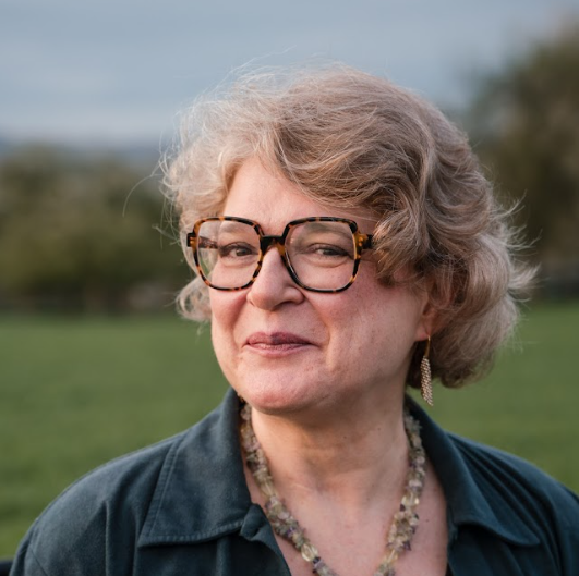 Anneke has short blonde hair and is wearing large tortoiseshell glasses. Anneke is stood in a field and is wearing a blue collared shirt and a statement necklace.