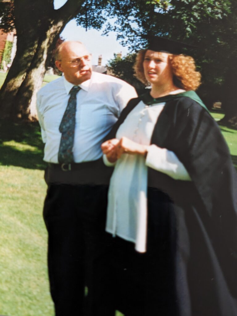 Helen is wearing a graduation cap and gown and is standing with a man wearing a suit and tie outside Ripon campus.